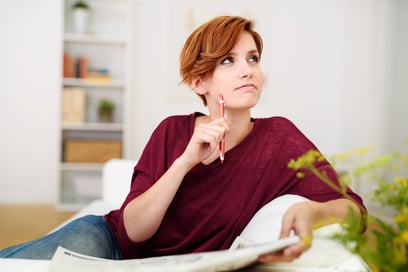 Thoughtful Attractive Young Woman Answering Crossword Puzzle Game on Newspaper at the Living Room Couch.